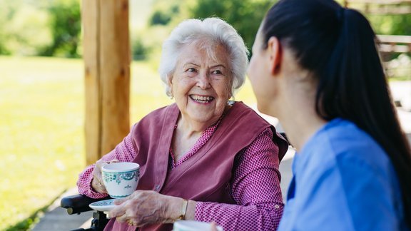 Female caregiver spending time with elderly woman, drinking coffee and talking. Nurse and senior woman in wheelchair enjoying a warm day outdoors.