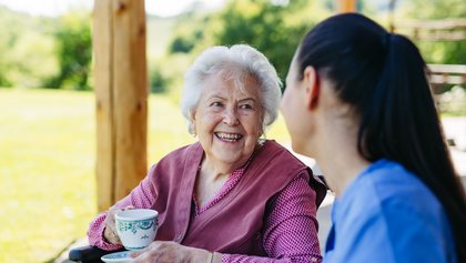 Female caregiver spending time with elderly woman, drinking coffee and talking. Nurse and senior woman in wheelchair enjoying a warm day outdoors.